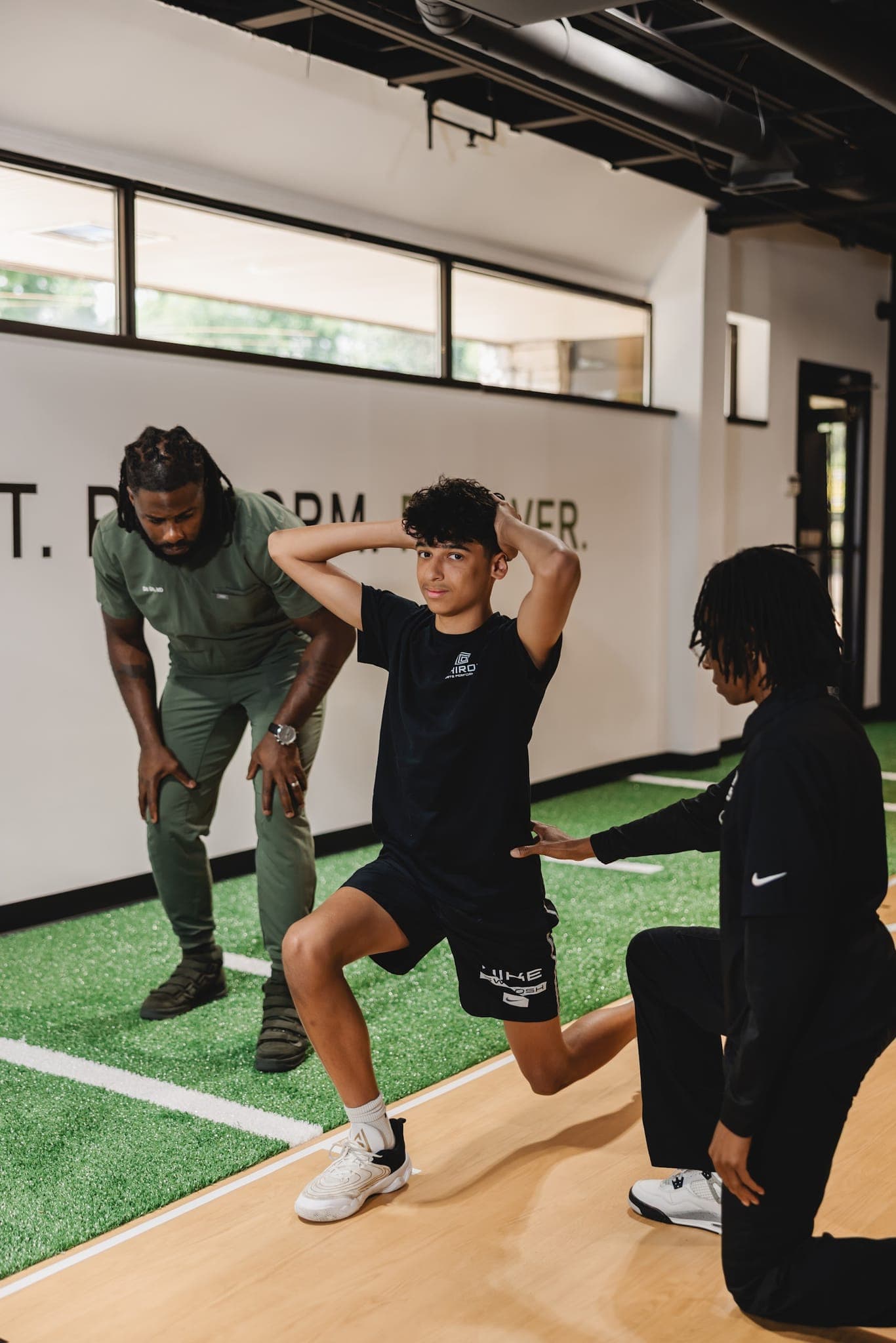A young athlete holds a lunge with his hands behind his head while one Chiron coach kneels beside him and another watches.