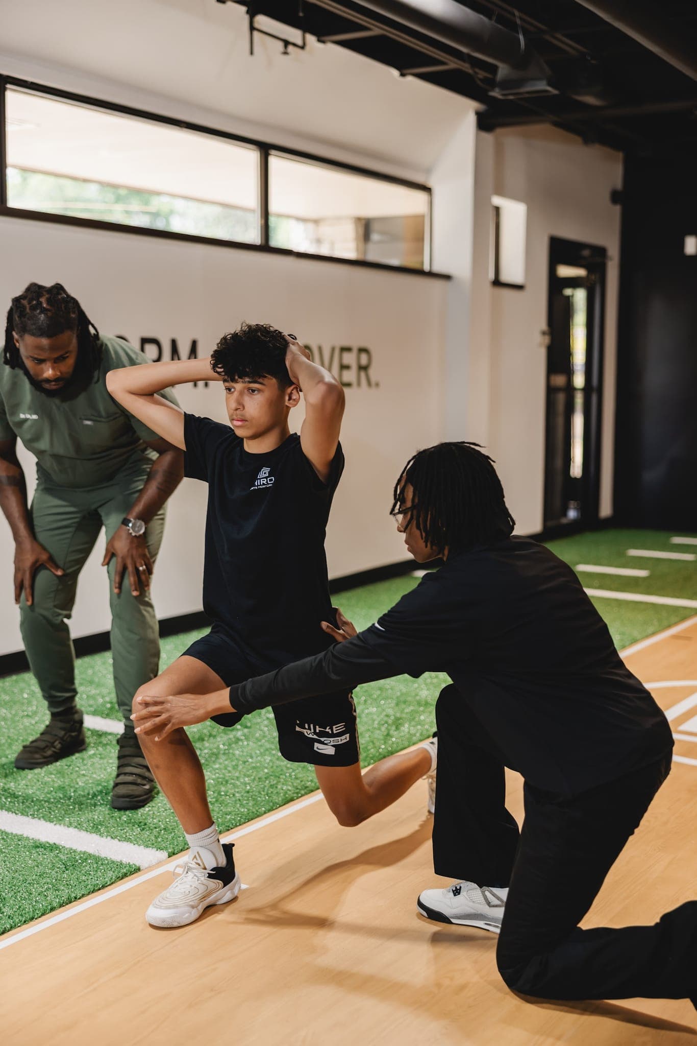 A Chiron coach kneels to adjust a young athlete's lunge, hands at his front knee, as a second coach looks on.