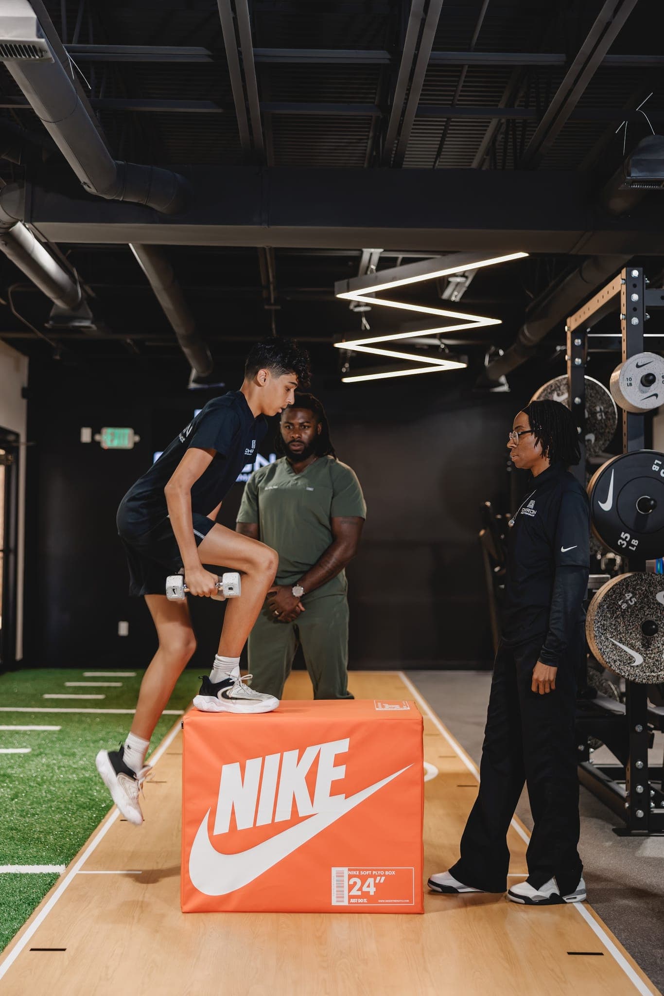 A young athlete steps up onto a 24-inch plyo box holding dumbbells as two Chiron coaches watch his form.