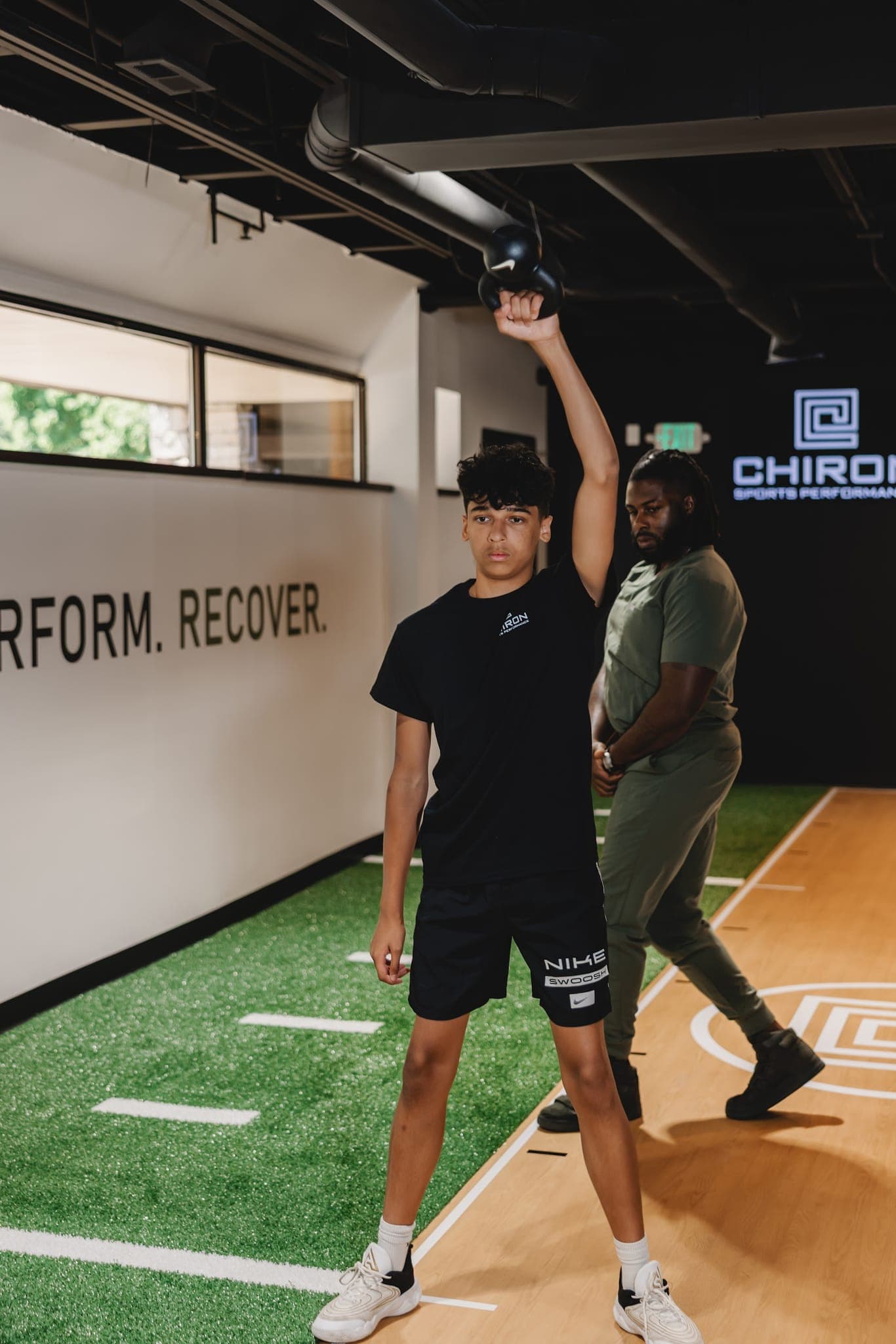 A young athlete holds a kettlebell overhead with one arm as a Chiron coach watches from behind, the Chiron Sports Performance sign lit up on the wall.