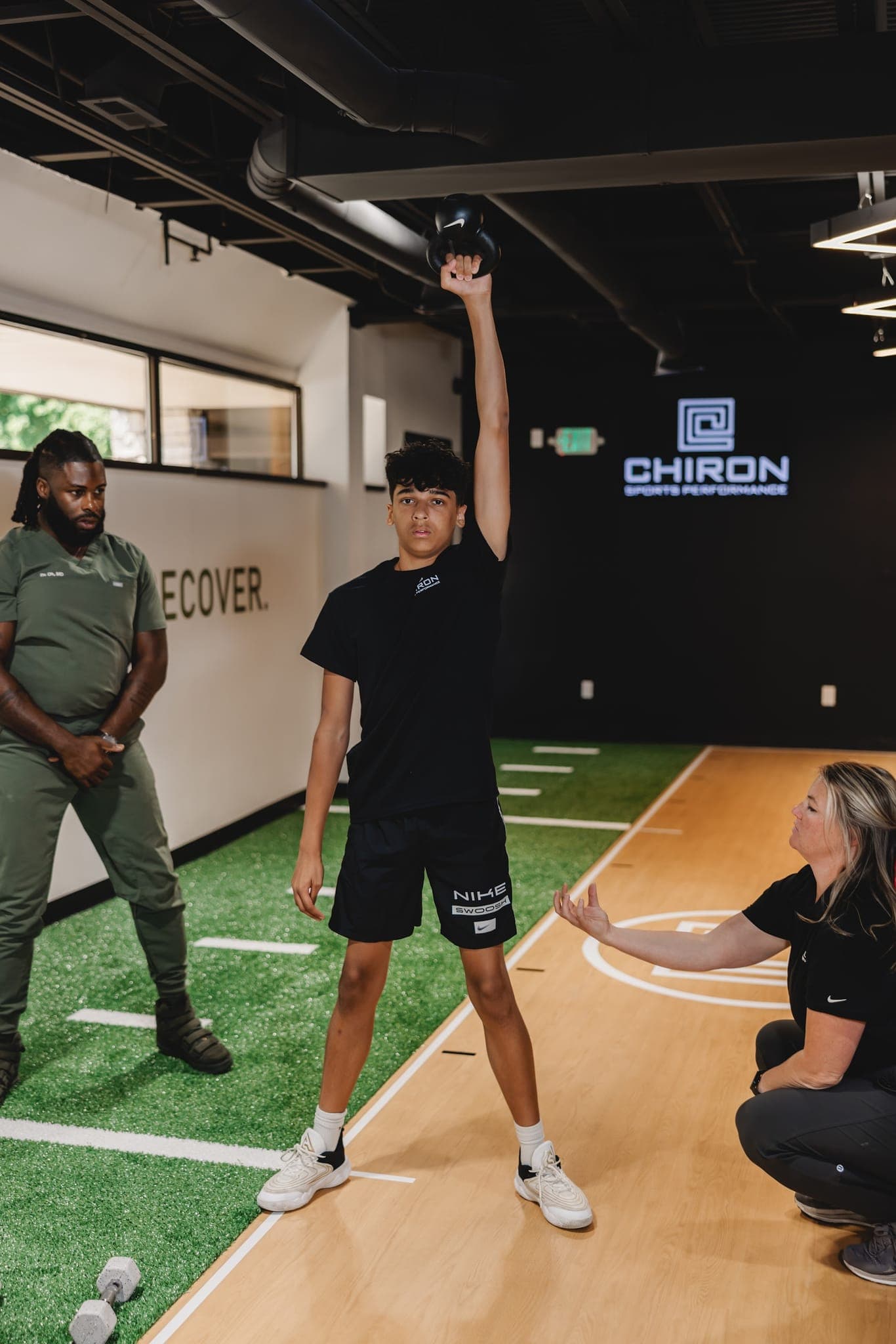 A Chiron coach crouches beside a young athlete holding a kettlebell overhead, coaching his posture, while another coach looks on.