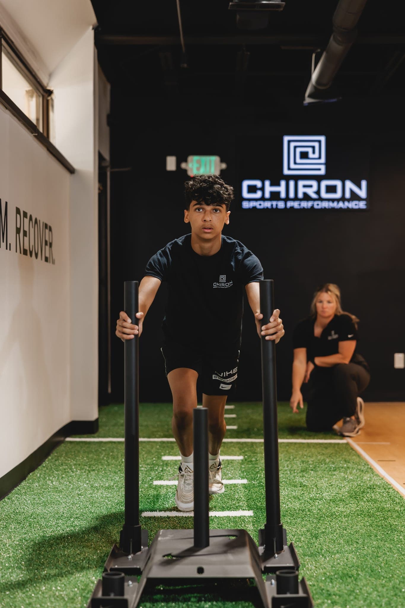 A young athlete drives a weighted sled across the turf toward the camera while a Chiron coach kneels and watches.