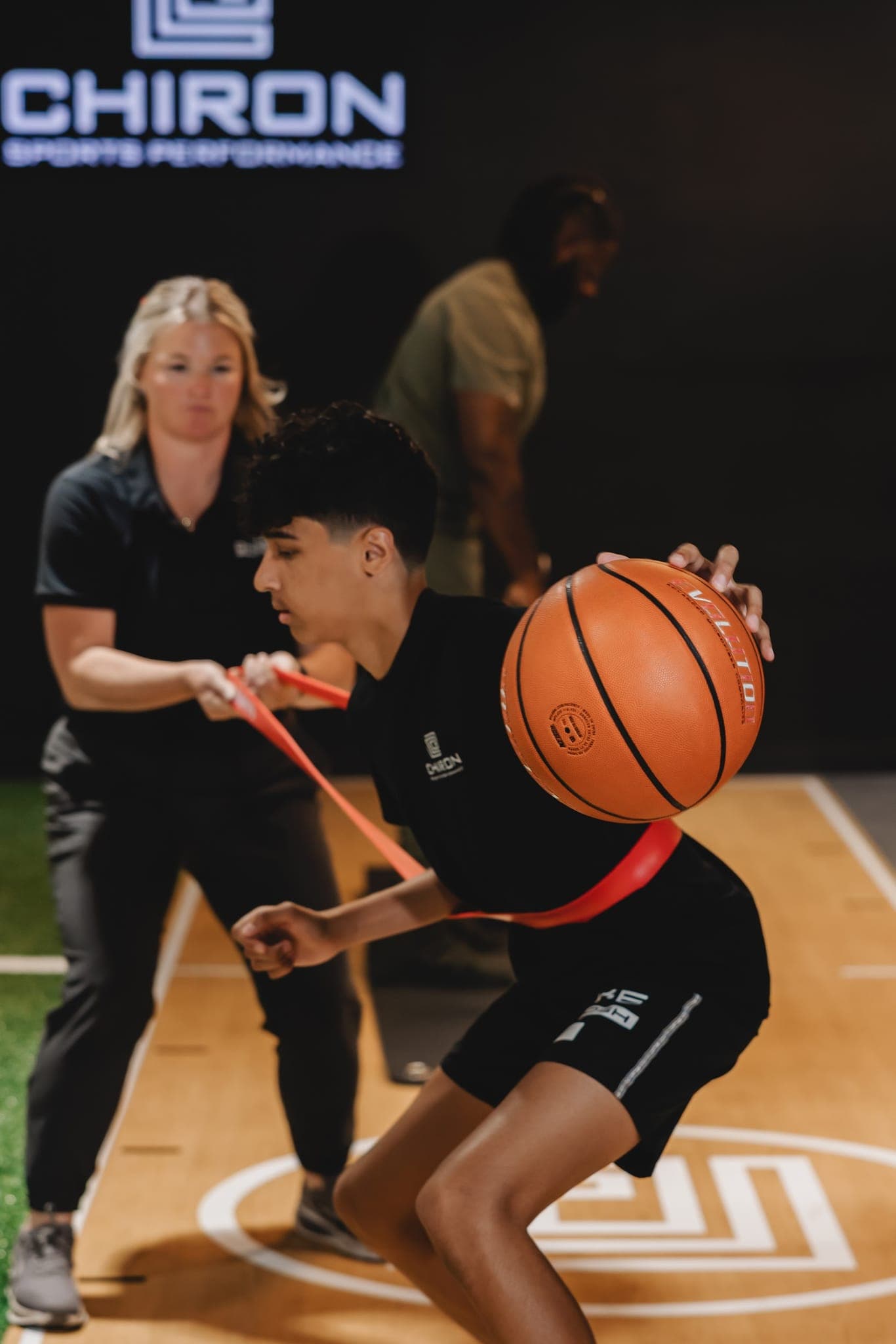 A young basketball player dribbles low against a red resistance band around his waist, held by a Chiron coach behind him.