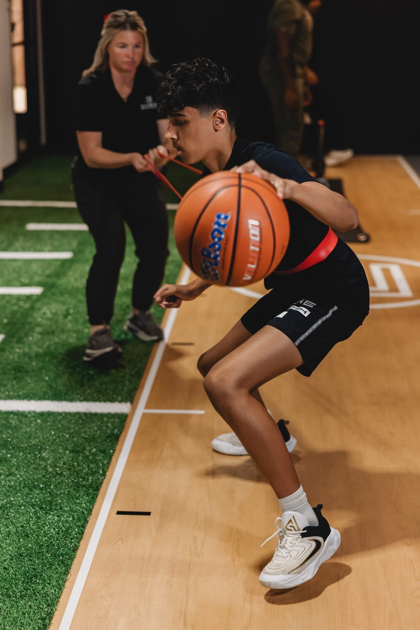A Chiron coach holds a resistance band around a young athlete's waist as he handles a basketball in a low athletic stance.