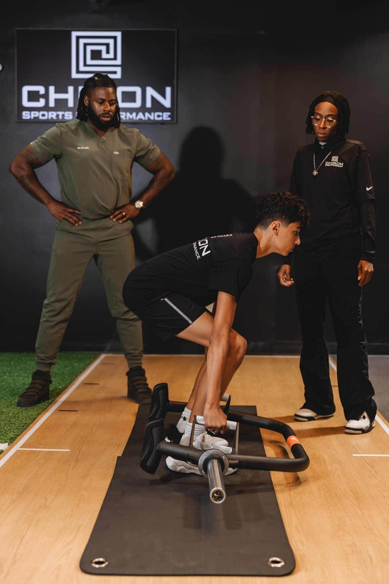 A young athlete sets up for a trap-bar deadlift on a mat while two Chiron coaches watch closely.