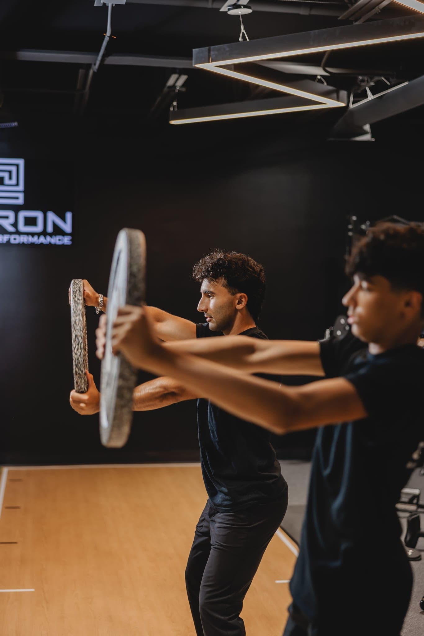 A Chiron coach and a young athlete press weight plates straight out at arm's length, side by side.